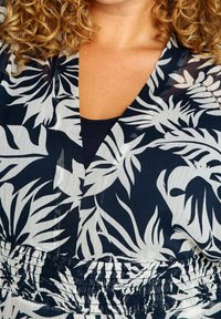 Person med lockigt hår iklädd en klänning med svart och vit tropisk lövmönster, med en samlad midja och ett svart inre lager synligt vid halsringningen.