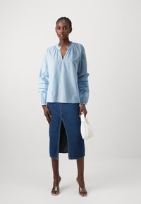 Blusa a righe blu chiaro con scollo a V e dettaglio arricciato, abbinata a una lunga gonna di jeans con uno spacco frontale e una clutch bianca.