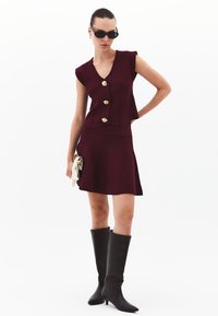 Burgundin mekko, jossa on V-pääntie, istuva yläosa ja kolme kultanappia. Yhdistetty polven korkeiden mustien saappaiden kanssa ja pidellen valkoista clutch-laukkua.