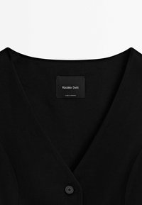Vêtement noir à col en V avec un seul bouton et une étiquette indiquant "Massimo Dutti, Fabriqué au Maroc" à l'intérieur de l'encolure.