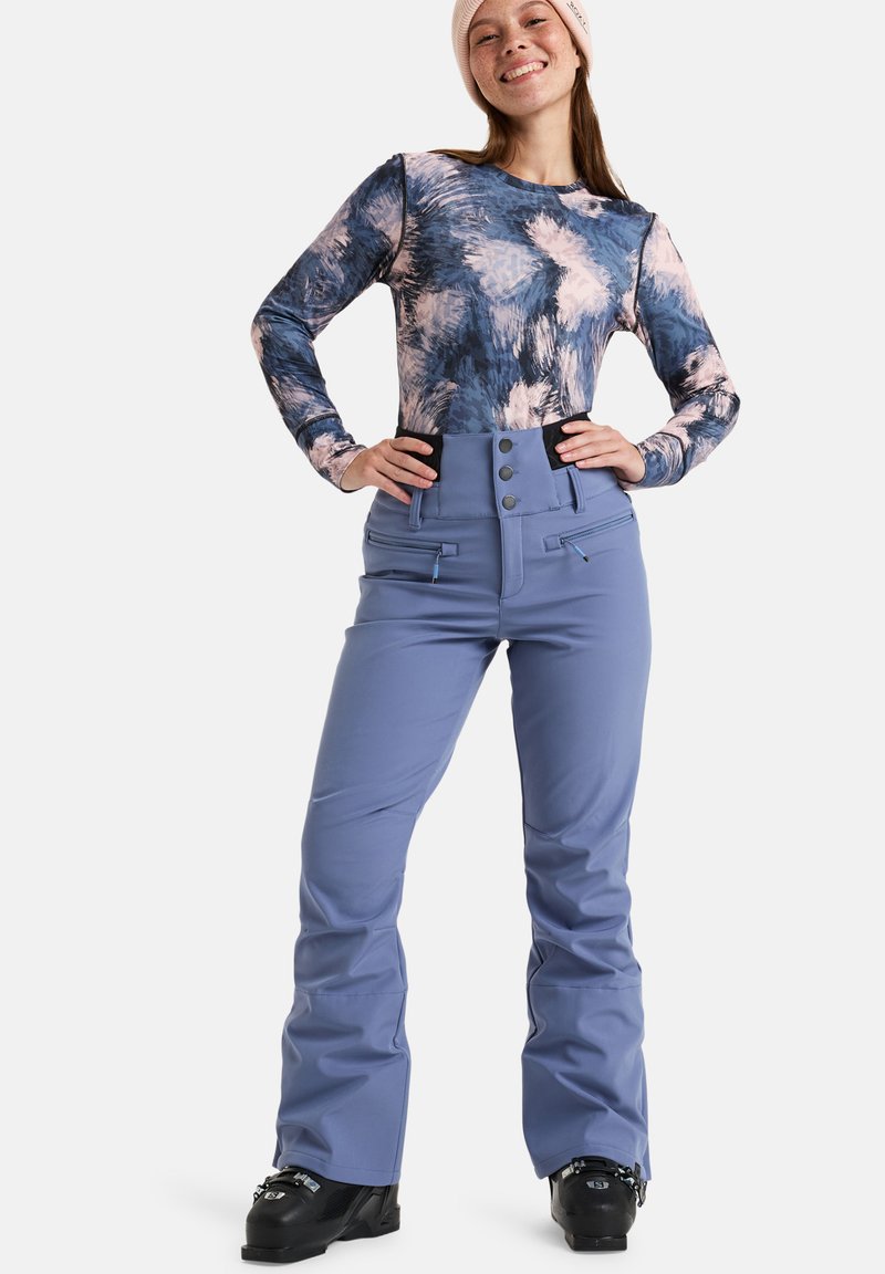 Blaue Skihosen mit hohem Bund, Knopfverschluss und Reißverschlusstaschen; kombiniert mit einem langärmligen, mehrfarbig gemusterten Oberteil.