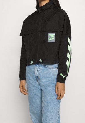 Personne portant une veste Puma noire avec un logo vert néon et des motifs sur la poitrine et la manche, associée à un jean bleu clair.