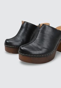 Zuecos de cuero negro sin cordones con dedos redondeados, costuras visibles y gruesos tacones plataforma de madera marrón sobre un fondo gris claro.