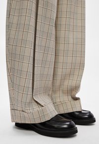 Calças largas de padrão xadrez bege com bainha virada, usadas com mocassins pretos. A textura parece suave e macia.