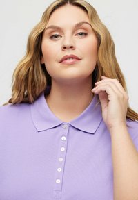 Liliowa koszulka polo z kołnierzykiem, wyposażona w listwę z pięcioma guzikami. Materiał ma miękką, teksturowaną powierzchnię.