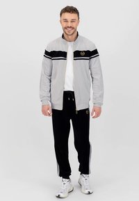 Grijze jas met ritssluiting, zwart gestreept detail en goudkleurig logo, gecombineerd met zwarte joggers met witte zijkstrepen en een trekkoord.