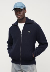 Marineblå strikket genser med glidelås og høy krage, med ribbestruktur, kombinert med en lyseblå jeanscaps og hvit t-skjorte under.