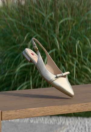 Chaussure plate à bride arrière métallique avec bout pointu et petit détail de nœud, posée sur une surface en bois avec un fond d'herbe verte.