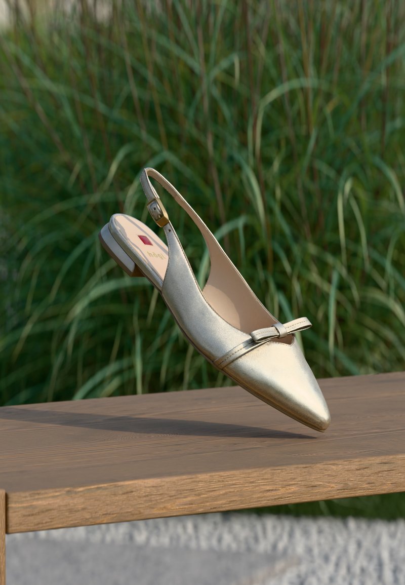 Chaussure plate à bride arrière métallique avec bout pointu et petit détail de nœud, posée sur une surface en bois avec un fond d'herbe verte.