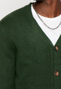 Donkergroene cardigan van gestructureerd gebreid materiaal, met een V-hals, drie knopen en een gelaagde witte blouse eronder.