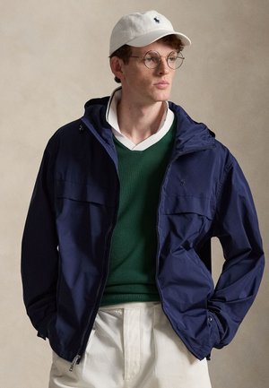 Ung mand iført runde briller, hvid kasket, navy bluse med lynlås, grøn sweater over hvid skjorte med krave og lyse bukser, kigger mod højre.