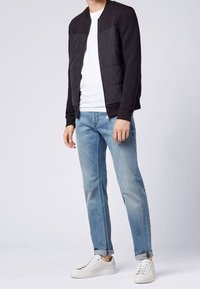 Hombre vistiendo una chaqueta bomber negra, camiseta blanca, jeans claros arremangados y zapatillas blancas, de pie contra un fondo sencillo.