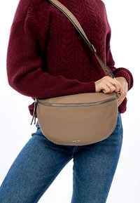 Křížová kožená taška světle hnědé barvy s kulatým tvarem, stříbrným zipem a béžovým popruhem. Nositelná s vínově červeným pleteným svetrem a modrými džíny.