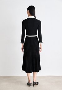 Robe noire plissée avec manches longues et col blanc. Elle présente un corsage ajusté et une jupe évasée, accompagnée de chaussures à talons noirs.