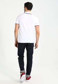 Maglietta polo bianca con un colletto scuro, abbinata a jeans scuri e scarpe da ginnastica rosse. Cuciture visibili e design classico.