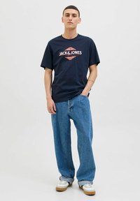 Jūrinės spalvos medvilnės marškinėliai su centriniu raudonu ir baltu grafinio logotipo dizainu, derinami su laisvais mėlynais džinsais ir baltais sportbačiais su pilkų akcentų.