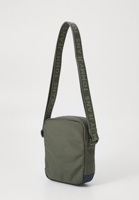 Olivově zelená crossbody taška s texturovanou tkaninou, námořnicky modrým dnem a vzorovaným popruhem, na kterém je tučným písmem napsáno "TOMMY JEANS".