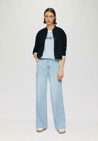 Veste de pilote navy, t-shirt rayé blanc et bleu avec texte, jeans larges en délavé clair, et chaussures à plateforme beiges. Tissu lisse, coupe décontractée.