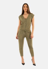 Tuta verde oliva con maniche corte, scollo a V, chiusura frontale con bottoni e vita con cordino; abbinata a tacchi alti neri.