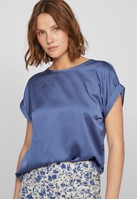 Blå sateng topp med korte ermer og rullede mansjetter, med en løs passform og rund halslinning. Stoffet har en glatt, skinnende tekstur.