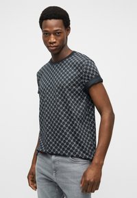 Νεαρός άνδρας που φοράει μαύρο πουκάμισο με σχέδια και κοντά μανίκια με γυρισμένα μανσόνια, καθώς και ανοιχτόχρωμα γκρι τζιν, στέκεται μπροστά σε απλό φωτεινό φόντο.