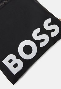 Czarna teksturowana saszetka z dużymi białymi literami "BOSS" nadrukowanymi diagonalnie na powierzchni i widocznym zamkiem błyskawicznym u góry.