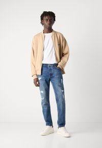 Beige bomber jacket, valkoinen t-paita, kuluneet siniset farkut ja valkoiset tennarit. Takissa on ribbireunat ja logo-osio.