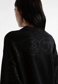 Μαύρο πλεκτό πουλόβερ με χαλαρή γραμμή, ριγέ λαιμό και διακριτό μοτίβο με κρανία στη πλάτη. Ο ιστός φαίνεται μαλακός και υφασματώδης.