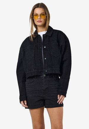 Melnā īsā denim jaka ar pogu priekšpusi, divām krūšu kabatām un brīvu piegriezumu, apvienota ar melniem denim šortiem. Dzeltenas saulesbrilles.