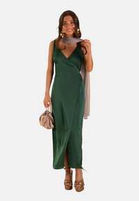 Vestido de satén verde con un escote pronunciado y una abertura alta. Combinado con una bufanda beige y un clutch topo, usado con tacones de plataforma.