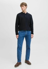 Joven de pie mirando a la izquierda, vistiendo una camiseta polo negra de manga larga, jeans azules, zapatos marrones, sobre un fondo claro y liso.