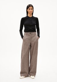 Schwarzes Langarm-Polohemd mit Knopfleiste, kombiniert mit weit geschnittenen braunen gemusterten Hosen. Das Model steht mit den Händen in den Taschen.