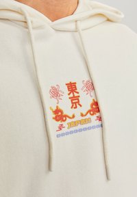 Närbild av en krämfärgad huvtröja med röda och gula Tokyo-kanji, två röda drakar och texten "Japan" i stiliserad font under tröjans snören.