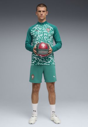 Uomo con completo sportivo verde del Portogallo tiene in mano un pallone da calcio rosso e verde del Portogallo, in piedi davanti a uno sfondo grigio.