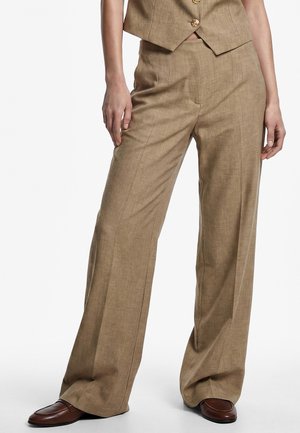 Kvinde iført beige bukser med vide ben og tilhørende vest samt brune læderloafers, står med hænderne afslappet ned langs siderne.