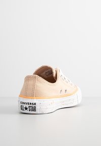 Baskets en toile beige à profil bas, semelle en caoutchouc blanc avec des accents orange et un motif tacheté. Logo "Converse All Star" sur le talon.