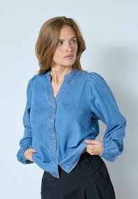 Blusa di denim in azzurro chiaro, con chiusura a bottone frontale, colletto alla mandarina e maniche a sbuffo. Tessuto liscio con polsini arricciati.