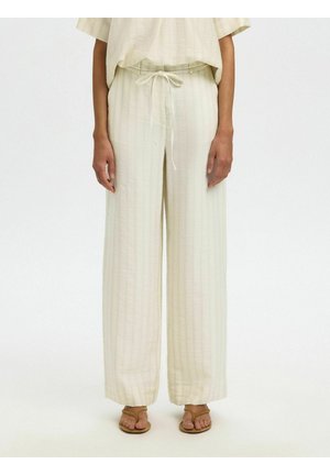 Person som har på seg løse, kremfargede bukser med høy midje, knappet midtlinje og subtile vertikale striper, kombinert med beige sandaler.