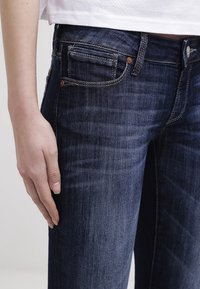 Jeans de mezclilla de color azul oscuro con corte slim, que cuentan con costuras en contraste y un diseño tradicional de cinco bolsillos. Textura suave con un ligero desgaste.