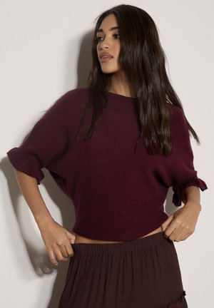Donna con capelli lunghi e scuri che indossa un maglione largo a maniche corte viola scuro e una gonna plissettata coordinata che si sistema la vita.