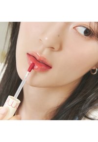 Produit à lèvres liquide avec une texture crémeuse dans un contenant présentant une base blanche et un texte rouge. L'applicateur est une brosse transparente et effilée.