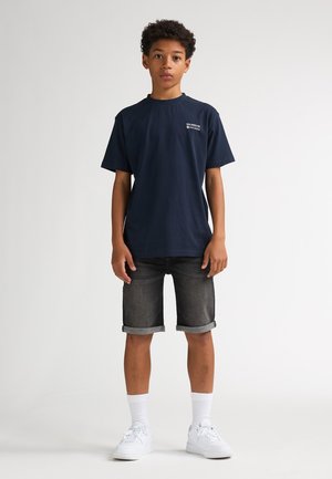 Tricou bleumarin cu decolteu rotund și mâneci scurte, asortat cu pantaloni scurți din denim închis la culoare. Se văd, de asemenea, adidași albi și șosete până la gleznă.