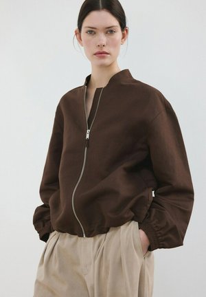 Femme portant une veste marron zippée avec des poignets élastiques et un pantalon plissé beige taille haute, debout devant un fond clair uni.