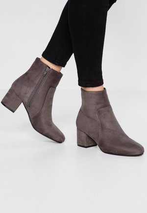Bottes chevilles en daim gris foncé avec talons carrés et fermetures éclair latérales portées avec un pantalon noir ajusté sur un sol blanc.