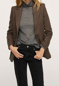 Blazer de cuadros marrones con un diseño estructurado, cierre de un solo botón, llevado sobre un suéter de cuello alto gris y combinado con jeans negros.