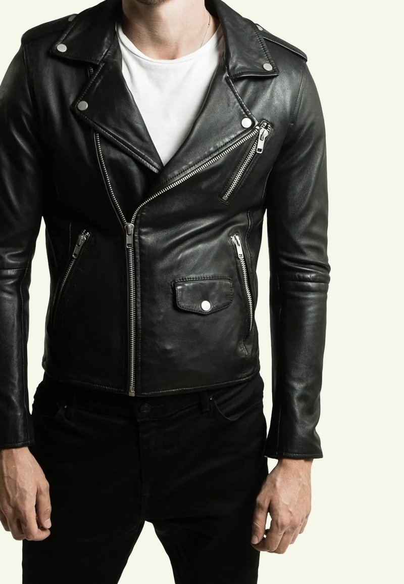 Homme portant une veste de motard en cuir noir avec des fermetures éclair et des boutons argentés, par-dessus un t-shirt blanc et un pantalon noir, debout les bras détendus.