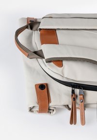 Gefalteter beiger Canvas-Rucksack mit braunen Lederakzenten, doppelten Reißverschluss-Zippern und Stoffgriffen vor weißem Hintergrund.
