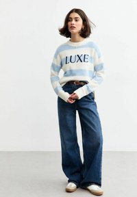 Maglione a righe blu e bianche con testo "LUXE", abbinato a jeans blu a vita alta e gamba larga e scarpe da ginnastica chiare. Texture morbide e vestibilità casual.