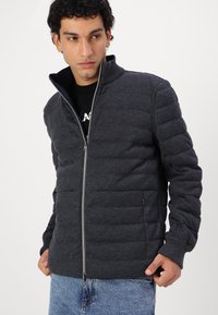 Veste rembourrée grise avec une fermeture éclair à l'avant, un col montant, des poignets côtelés et des poches latérales, associée à un t-shirt noir et un jean bleu déchiré.