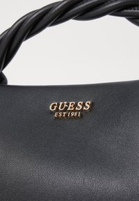 Black synthetic leather handbag with a twisted handle, featuring a gold "GUESS EST 1981" logo. Smooth texture with subtle detailing.